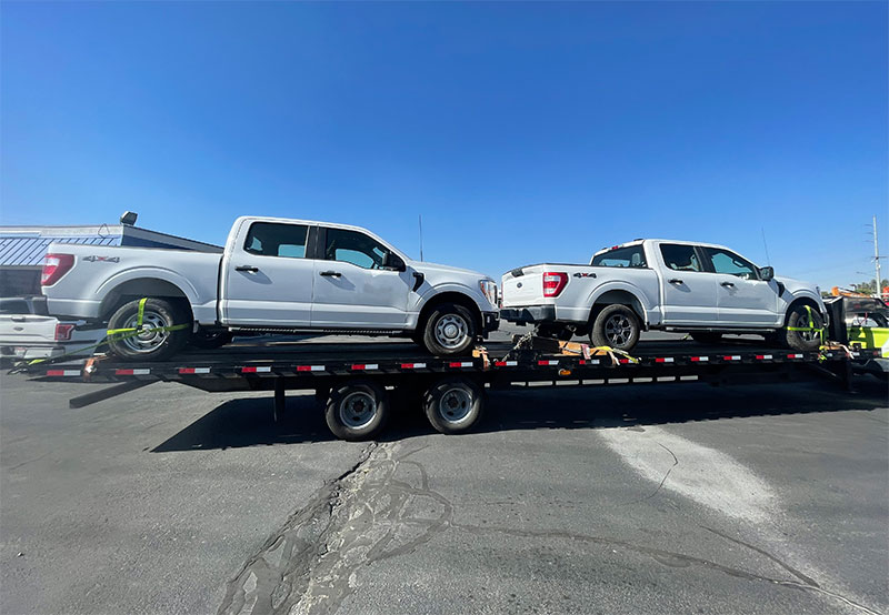 Two white four-door trucks on a flatbed that is parked in front of a blue paneled building.