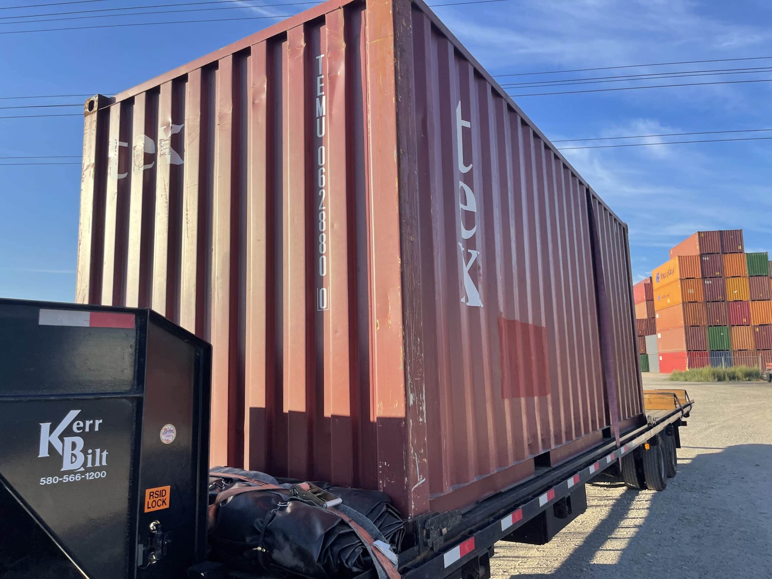 A red cargo worthy shipping container on a flatbed truck. The truck has "Kerr Built 580-566-1200" on the side and multi-colored shipping containers behind it.