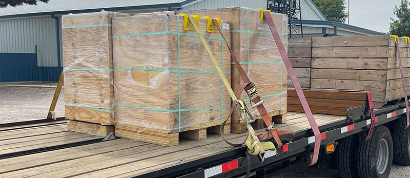 A flatbed truck with several packages of wood strapped to the truck. The truck is in front of a gray and blue paneled building.